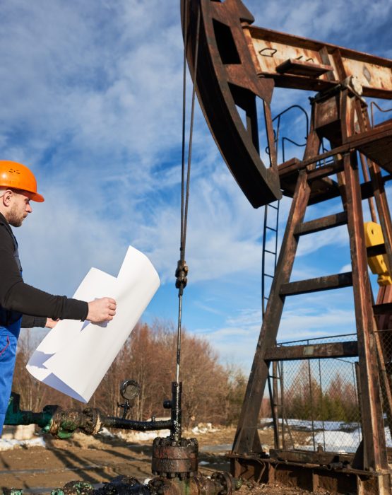 Side view of oil worker holding plan of oil field. Engineer in work uniform and helmet standing near petroleum pump jack under beautiful cloudy sky. Concept of petroleum industry and oil extraction.